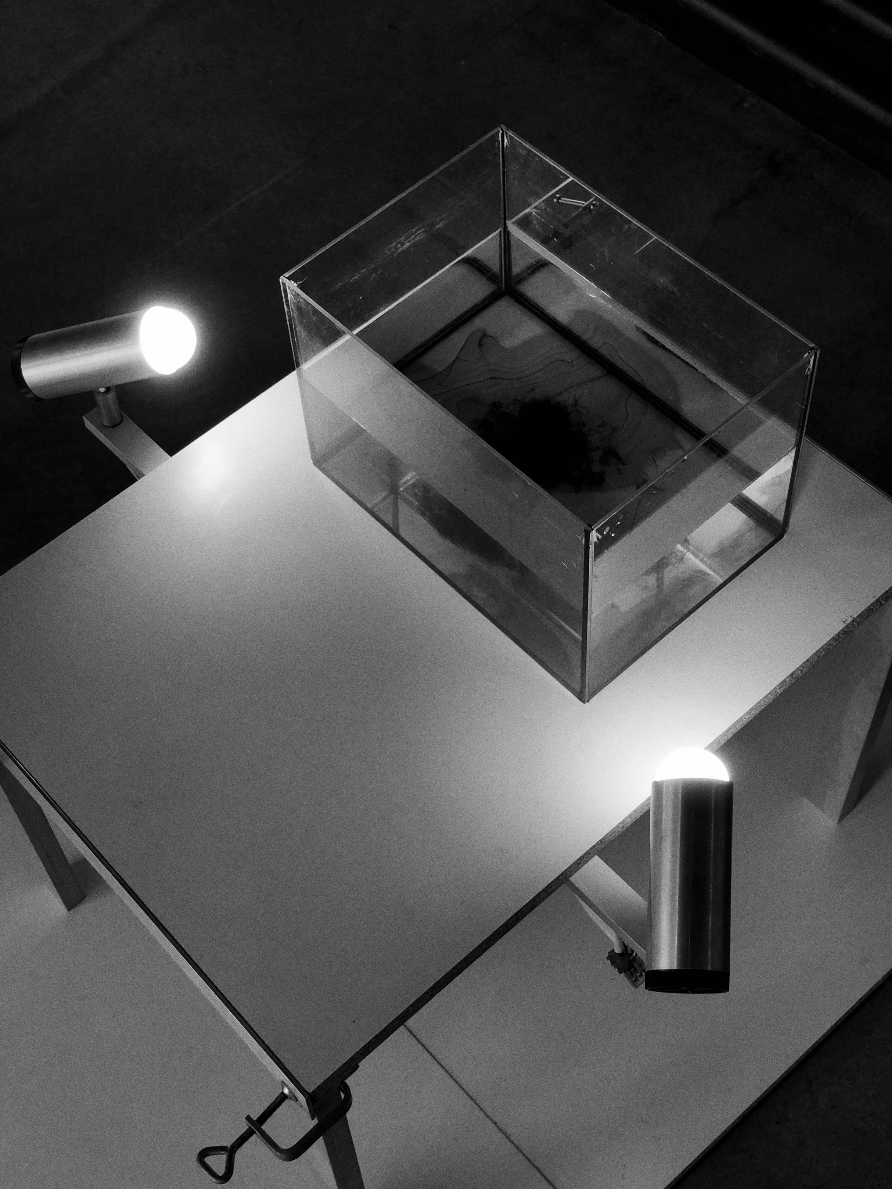 Black and white photo of a glass tank with a drop of ink in water sitting on a clamped-together table. Two spotlights